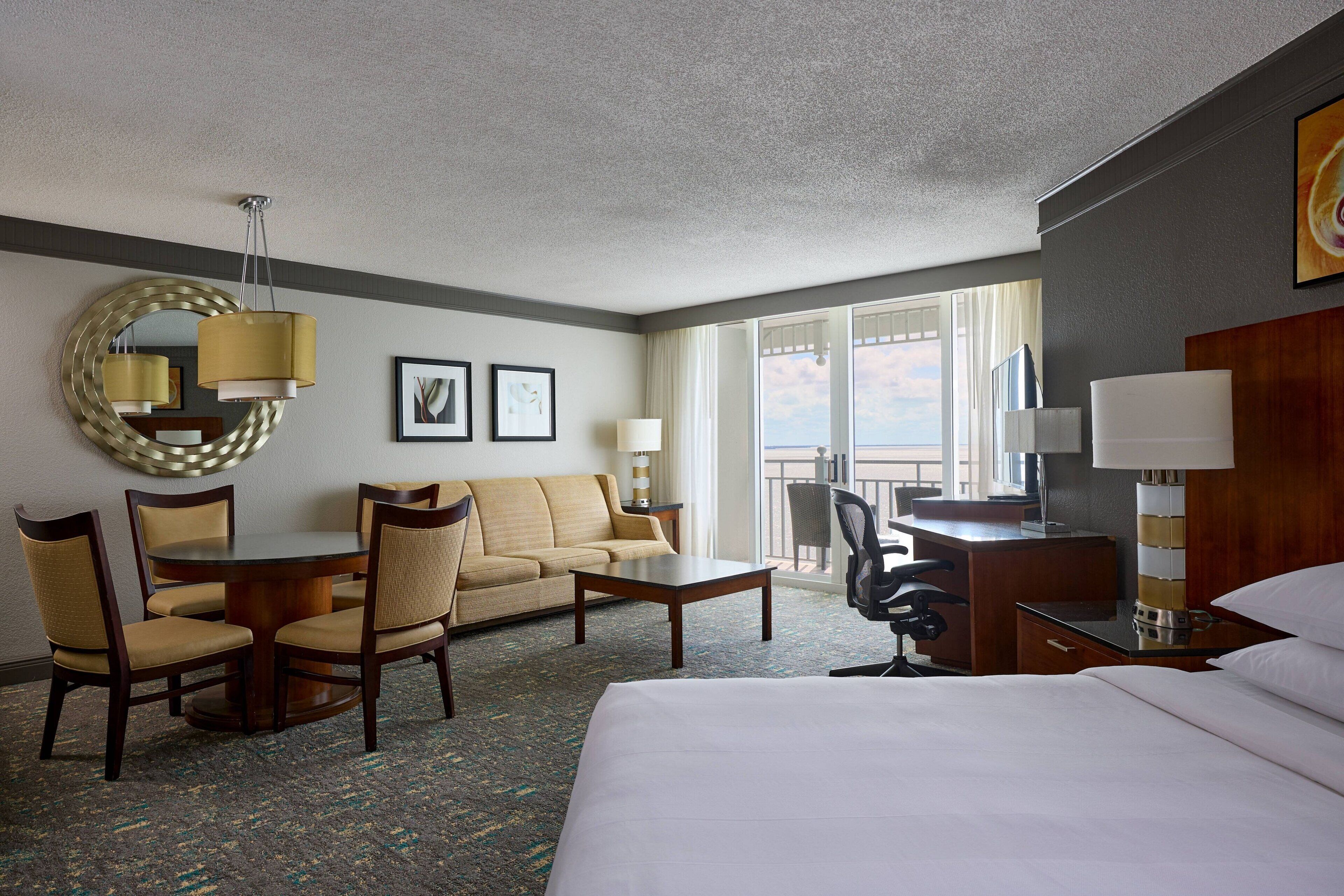 A hotel room with a bed, a round dining table with chairs, a sofa, a desk with a chair, a lamp, and a large window with a view.