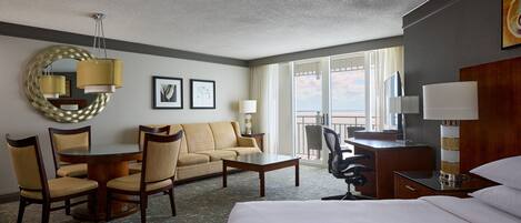 A hotel room with a bed, a round dining table with chairs, a sofa, a desk with a chair, a lamp, and a large window with a view.
