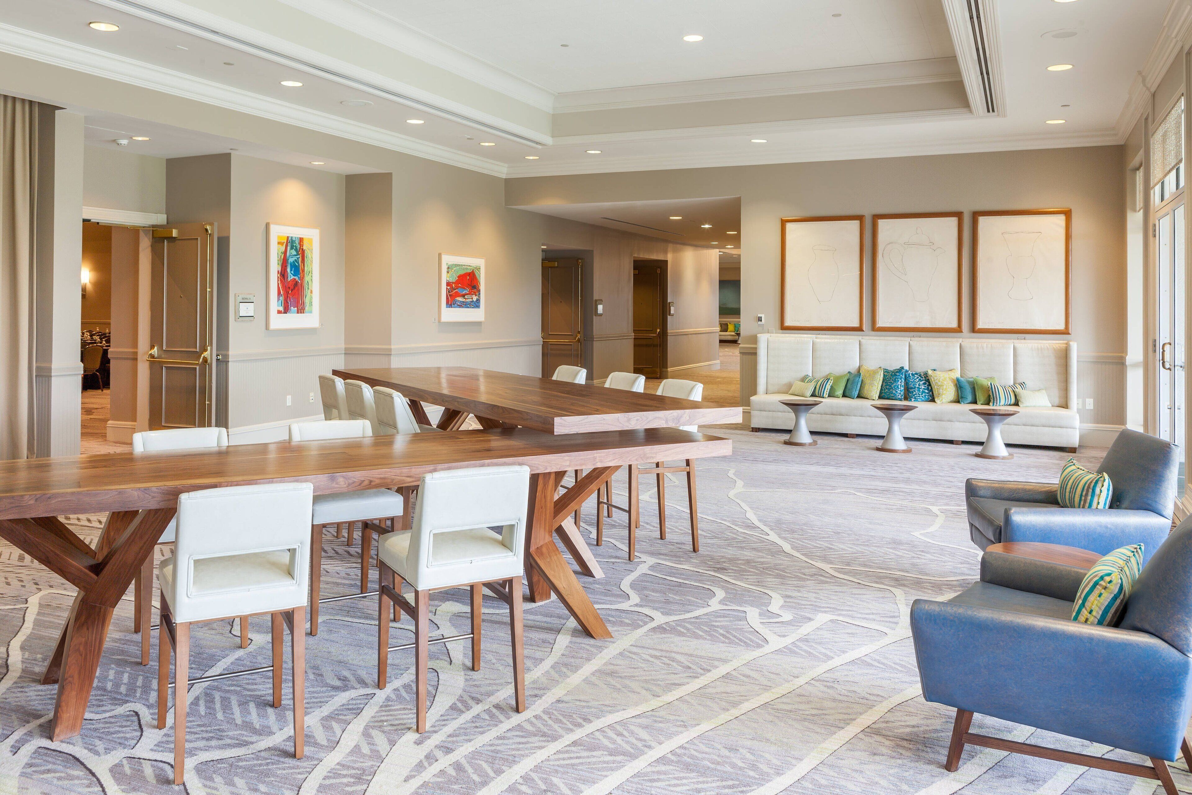 A spacious hotel lobby with a large wooden conference table, white chairs, a grey sofa, and a patterned carpet.
