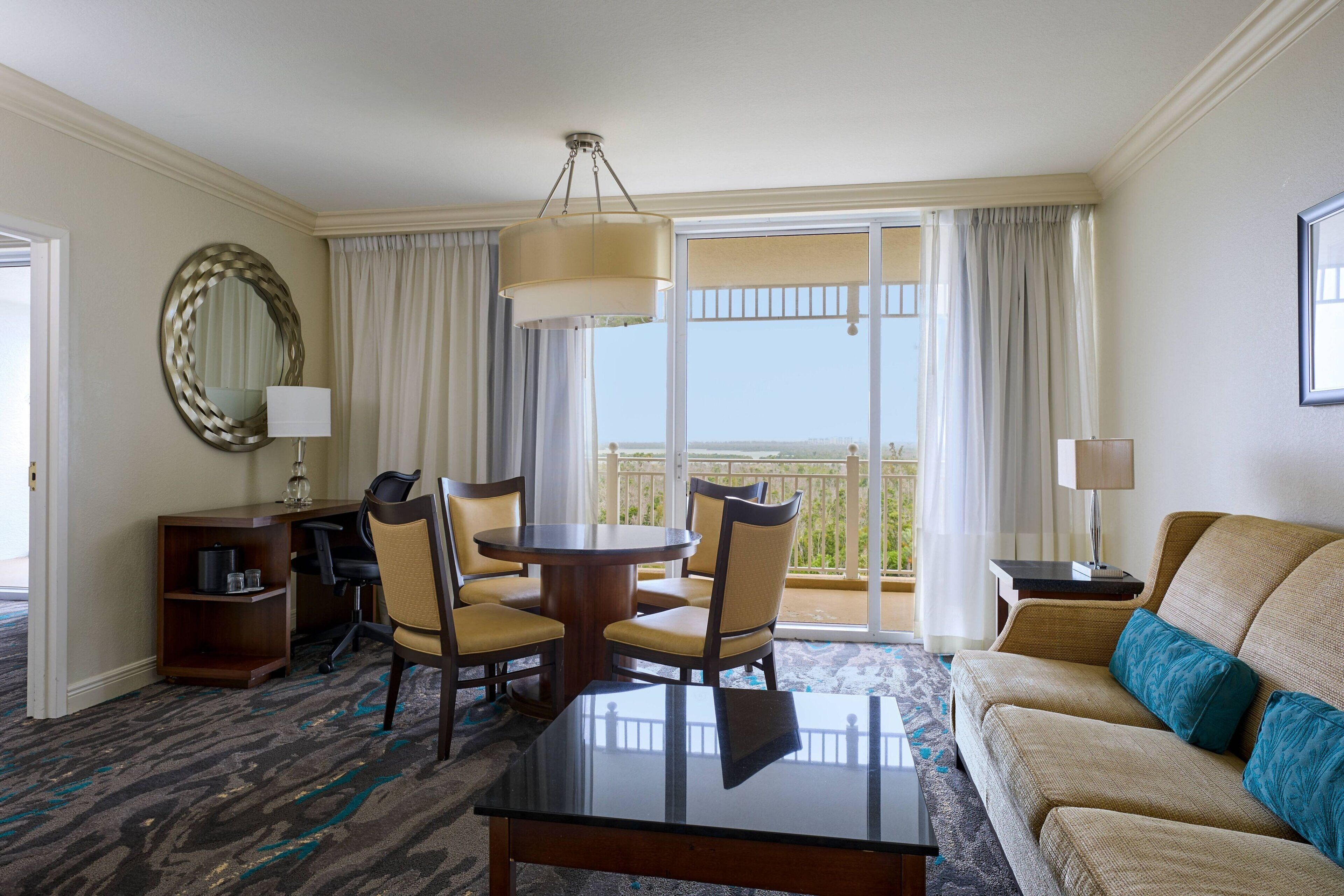 A hotel room with a balcony, a dining table with chairs, a sofa, and a desk with a lamp.