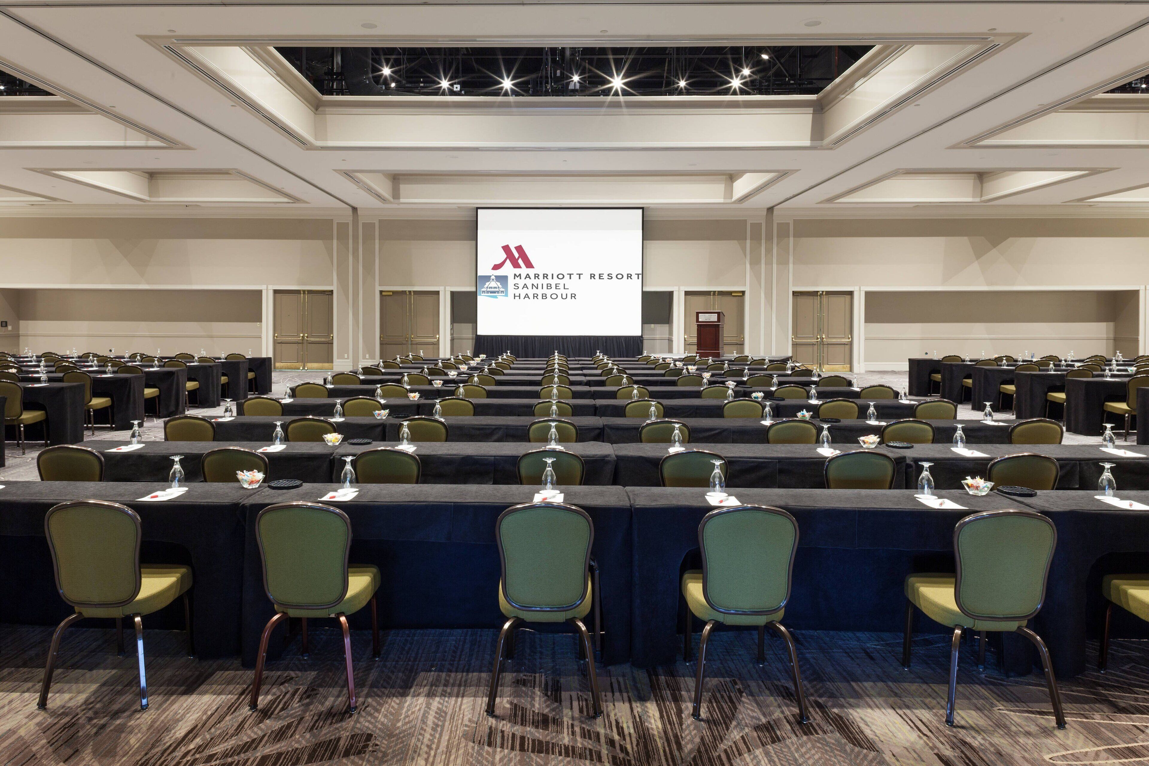 Ein großer Konferenzraum mit Tischen und Stühlen in Reihen, einem Bildschirm mit dem Marriott-Logo und einer Bühne.