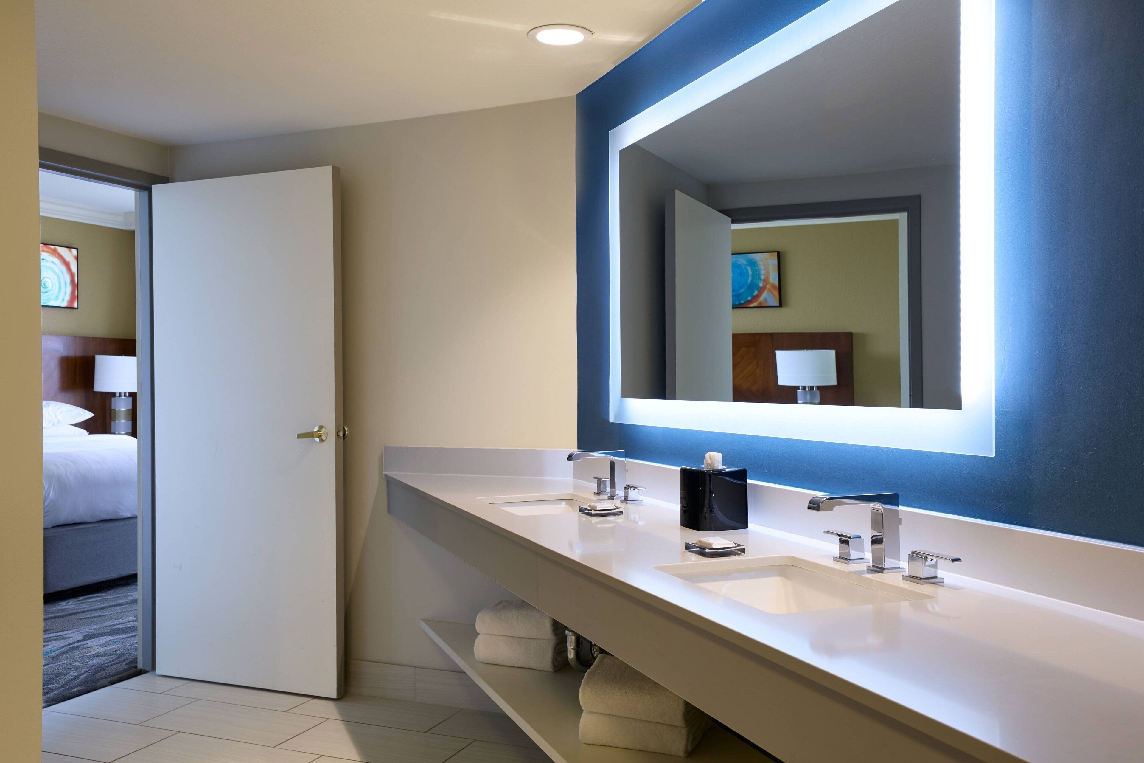 Une salle de bain moderne d’hôtel, dotée d’un grand miroir, de deux vasques et d’une porte donnant sur une chambre.