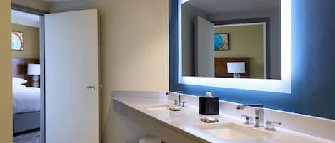A modern hotel bathroom with a large mirror, two sinks, and a door leading to a bedroom.