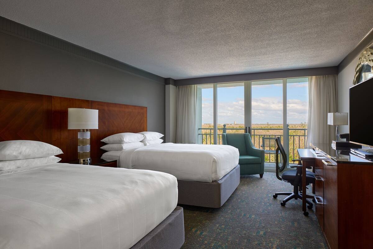 A hotel room with two beds, a desk, a chair, and a large window with a view.