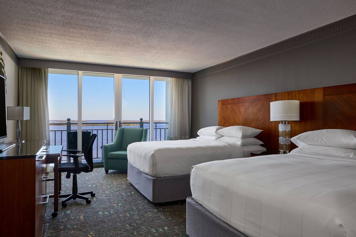 A hotel room with two beds, a desk, a chair, and a view of the sea.