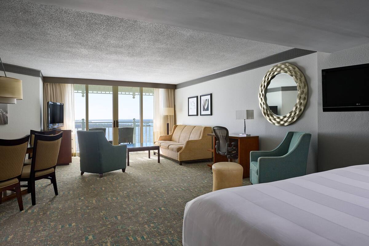 A hotel room with a large bed, a sofa, a desk with a computer, a TV, and a view of the sea through large windows.