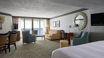 A hotel room with a large bed, a sofa, a desk with a computer, a TV, and a view of the sea through large windows.