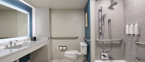 Un baño moderno con lavamanos, inodoro y bañera blancos. Incluye un cabezal de ducha y barras de apoyo.