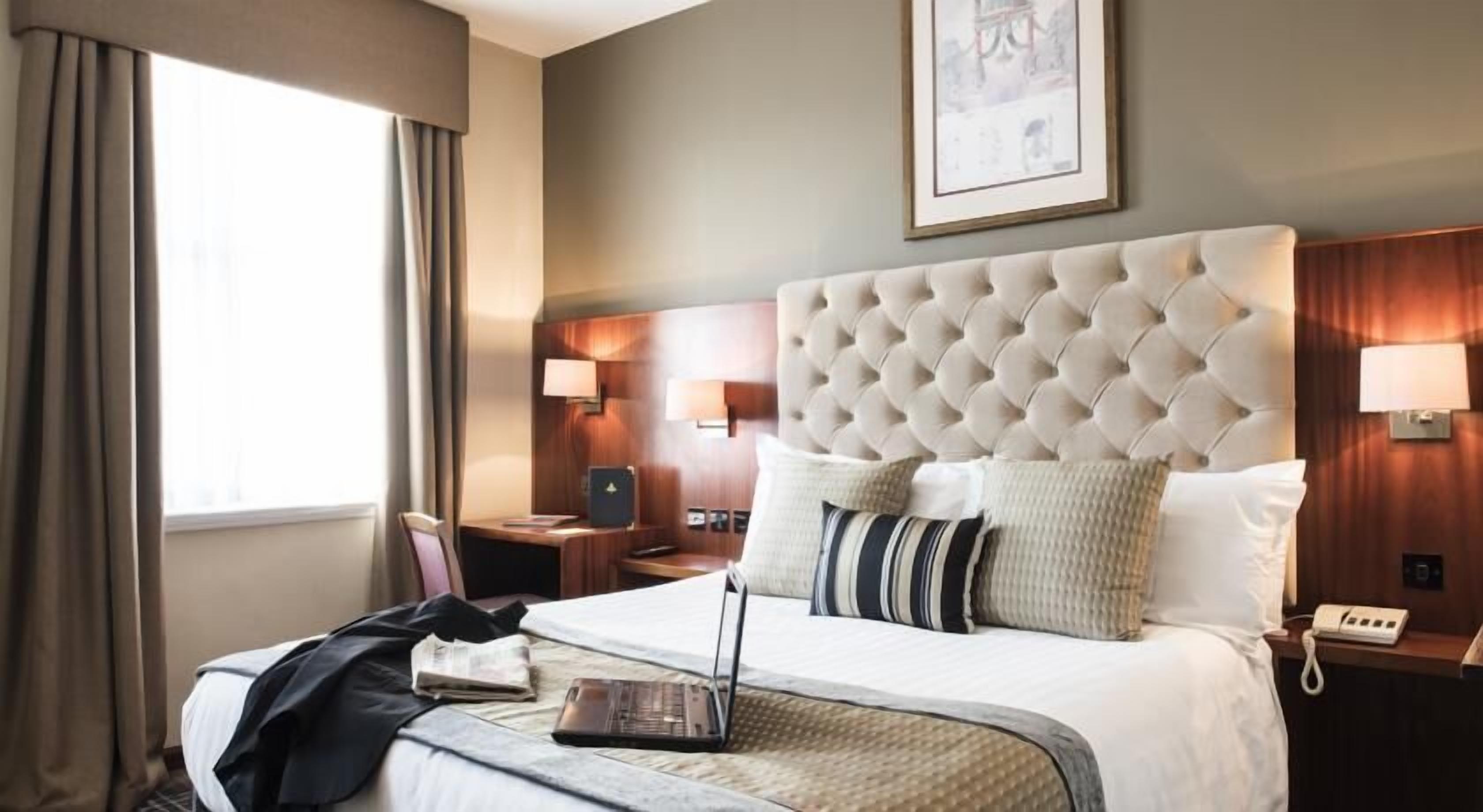 A hotel room with a large bed, a desk, a chair, a laptop, a telephone, and a framed picture on the wall.