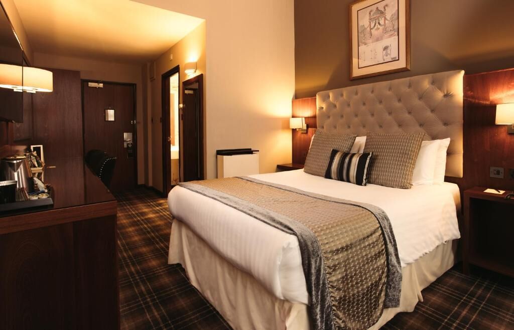 A hotel room with a large bed, a desk, and a chair. There are two bedside lamps and a framed picture on the wall.