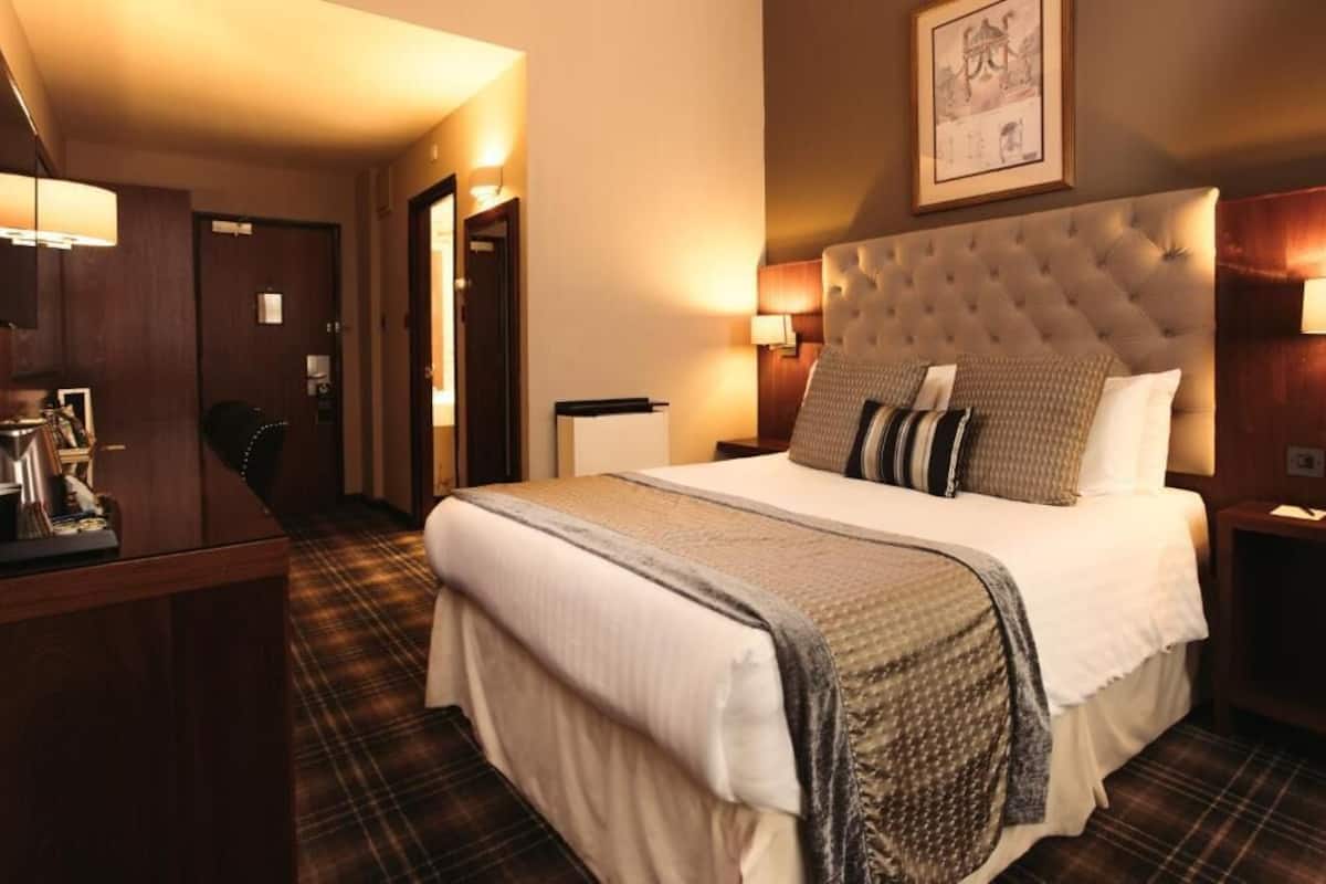 A hotel room with a large bed, a desk with a lamp, and a door leading to another room.