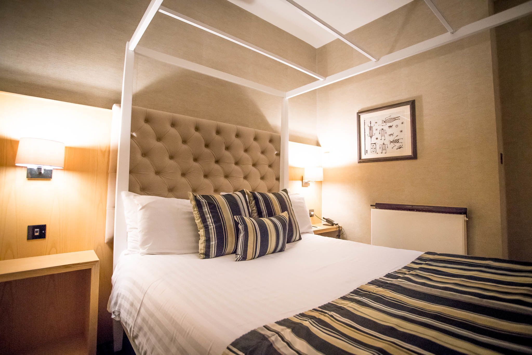 A hotel room with a four-poster bed, a wooden nightstand, a bedside table, and a wall-mounted lamp.