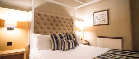A hotel room with a four-poster bed, a wooden nightstand, a bedside table, and a wall-mounted lamp.