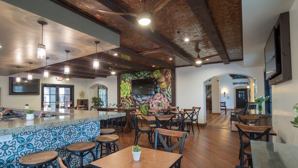 Un restaurante espacioso con techo alto, vigas de madera y un mural colorido. Hay varias mesas y sillas, y se observa una estación de café al fondo.