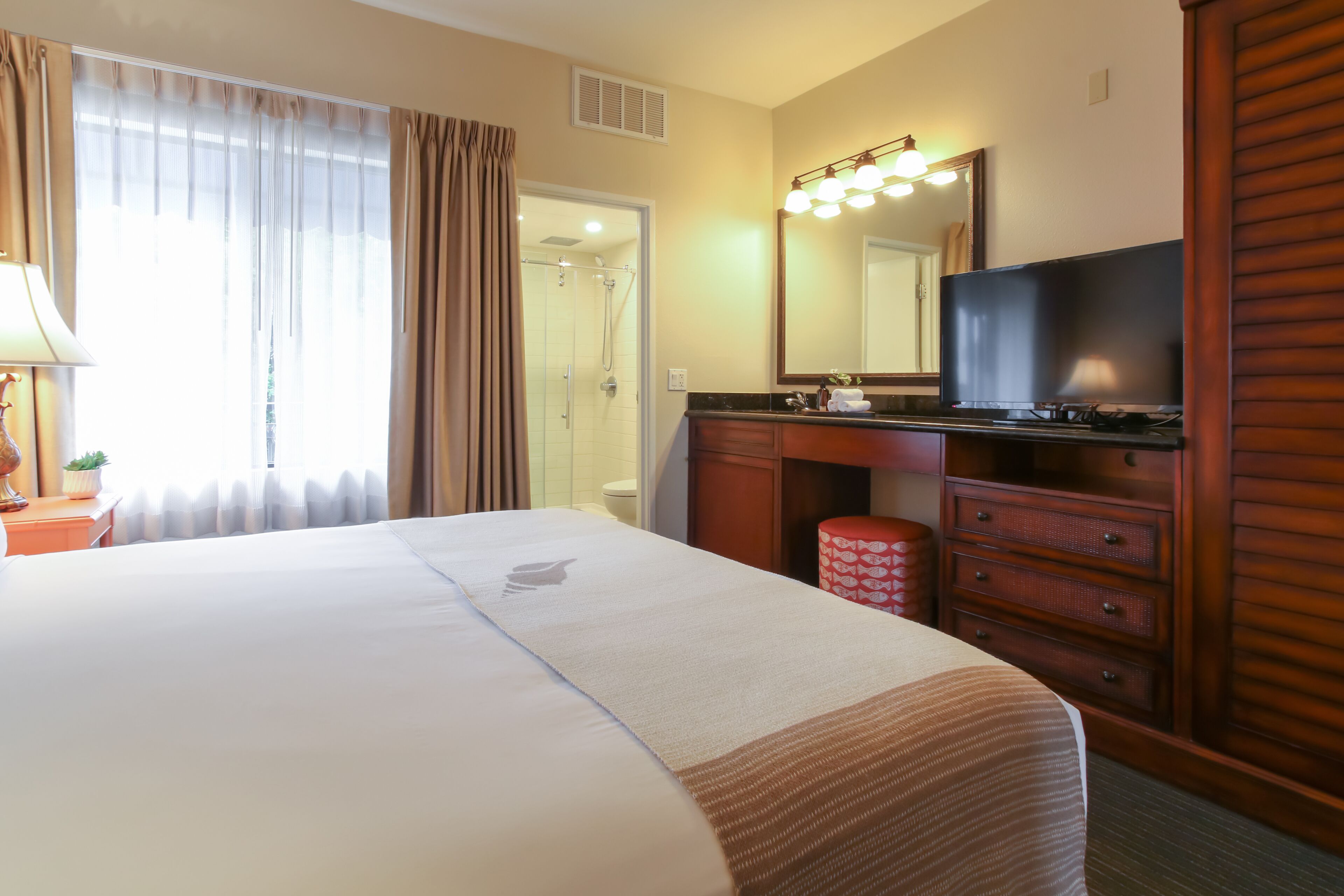 A hotel room with a large bed, a bathroom with a shower, a vanity with a mirror, and a television.