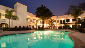 Una piscina con sillas de descanso, palmeras y un edificio con varios balcones e iluminación.