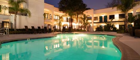 A swimming pool with lounge chairs, palm trees, and a building with multiple balconies and lights.