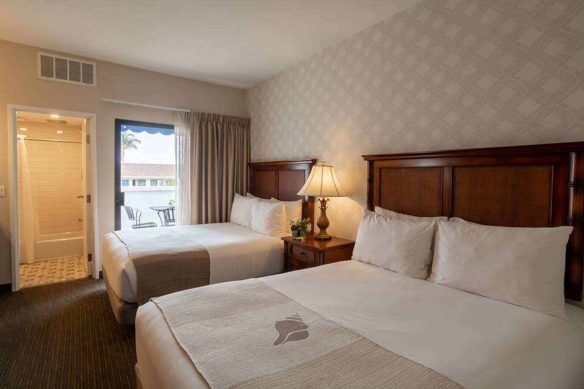 Een hotelkamer met twee bedden, een nachtkastje met een lamp en uitzicht op het omliggende landschap door een raam.