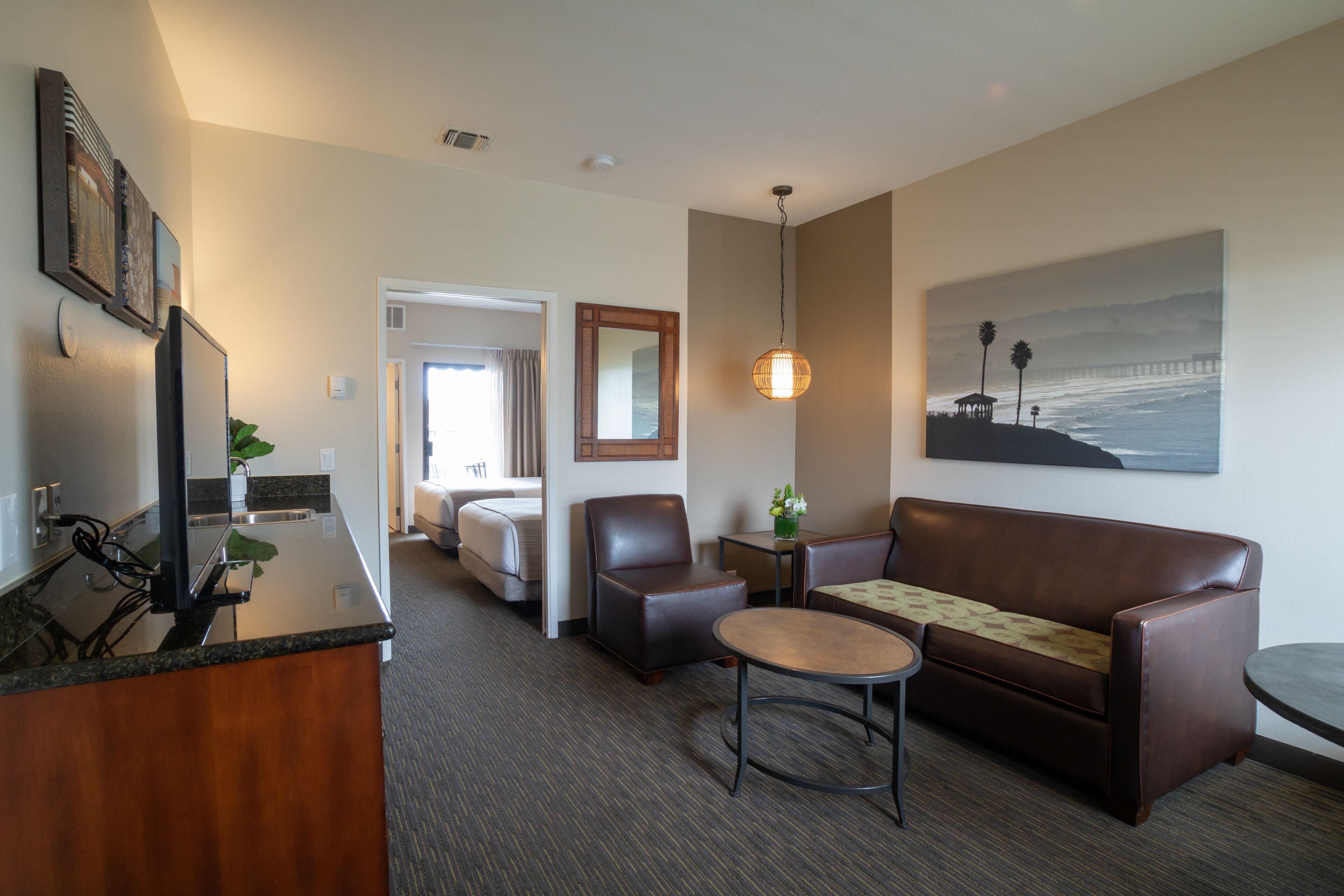A hotel room with a kitchenette, a sofa, a coffee table, a desk, and a painting of a beach scene.