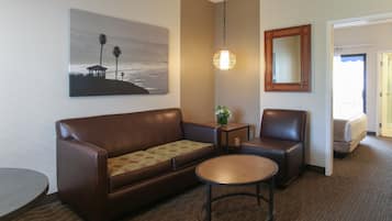 Ein Hotelzimmer mit einer braunen Ledersofa, einem runden Couchtisch, einem kleinen Beistelltisch mit einer Pflanze, einem großen Spiegel und einem Gemälde mit Strandmotiv.