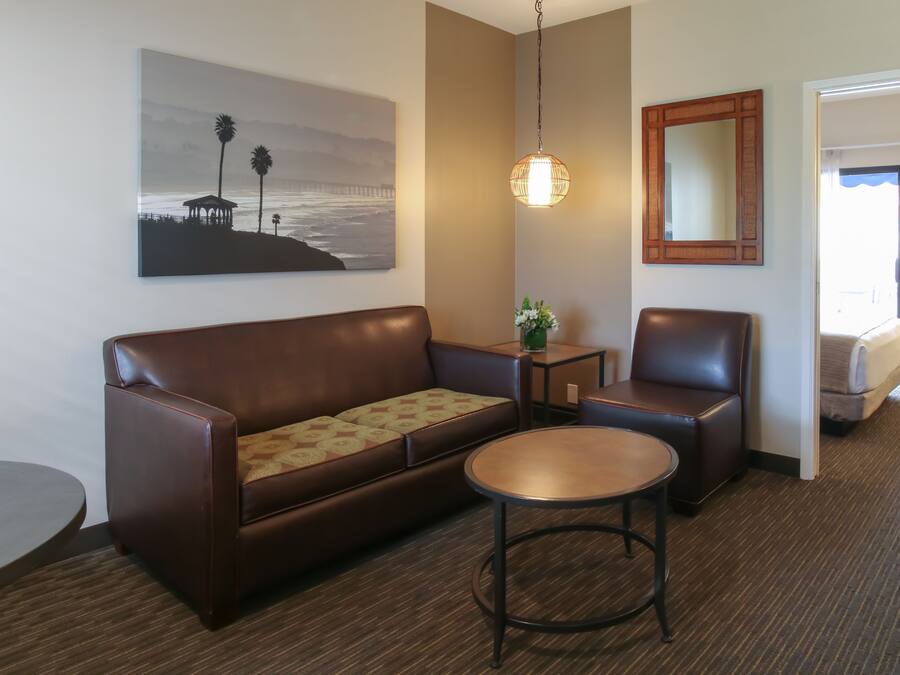 Ein Hotelzimmer mit einer braunen Ledersofa, einem runden Couchtisch, einem kleinen Beistelltisch mit einer Pflanze, einem großen Spiegel und einem Gemälde mit Strandmotiv.