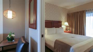 A hotel room with a large bed, a wooden headboard, a bedside table with a lamp, and a small desk with a chair and a vase of flowers.