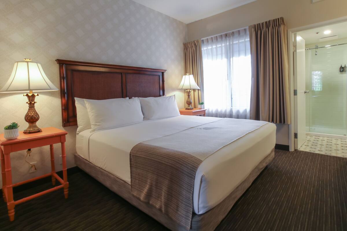 Hotelkamer met een groot bed, twee nachtkastjes met lampen, een kleine potplant en een glazen douchedeur.