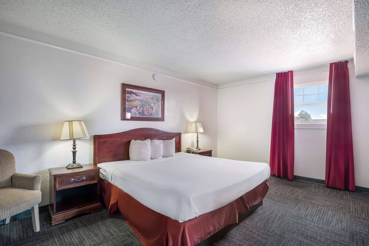 A hotel room with a large bed, two bedside tables with lamps, a chair, and a window with red curtains.