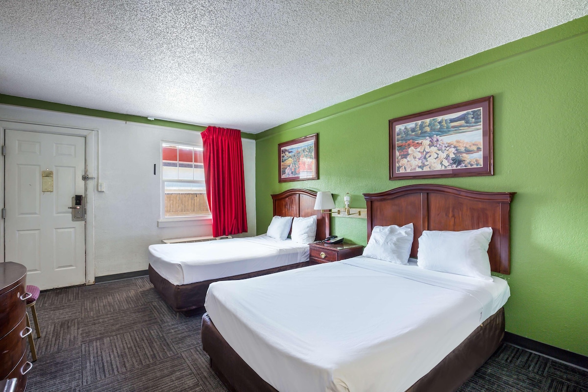 A hotel room with two beds, a desk, a chair, a window with red curtains, and two paintings on the wall.