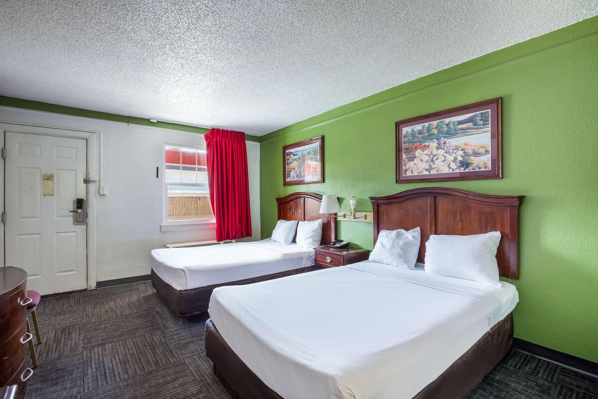 A hotel room with two beds, a desk, a chair, a window with red curtains, and two paintings on the wall.