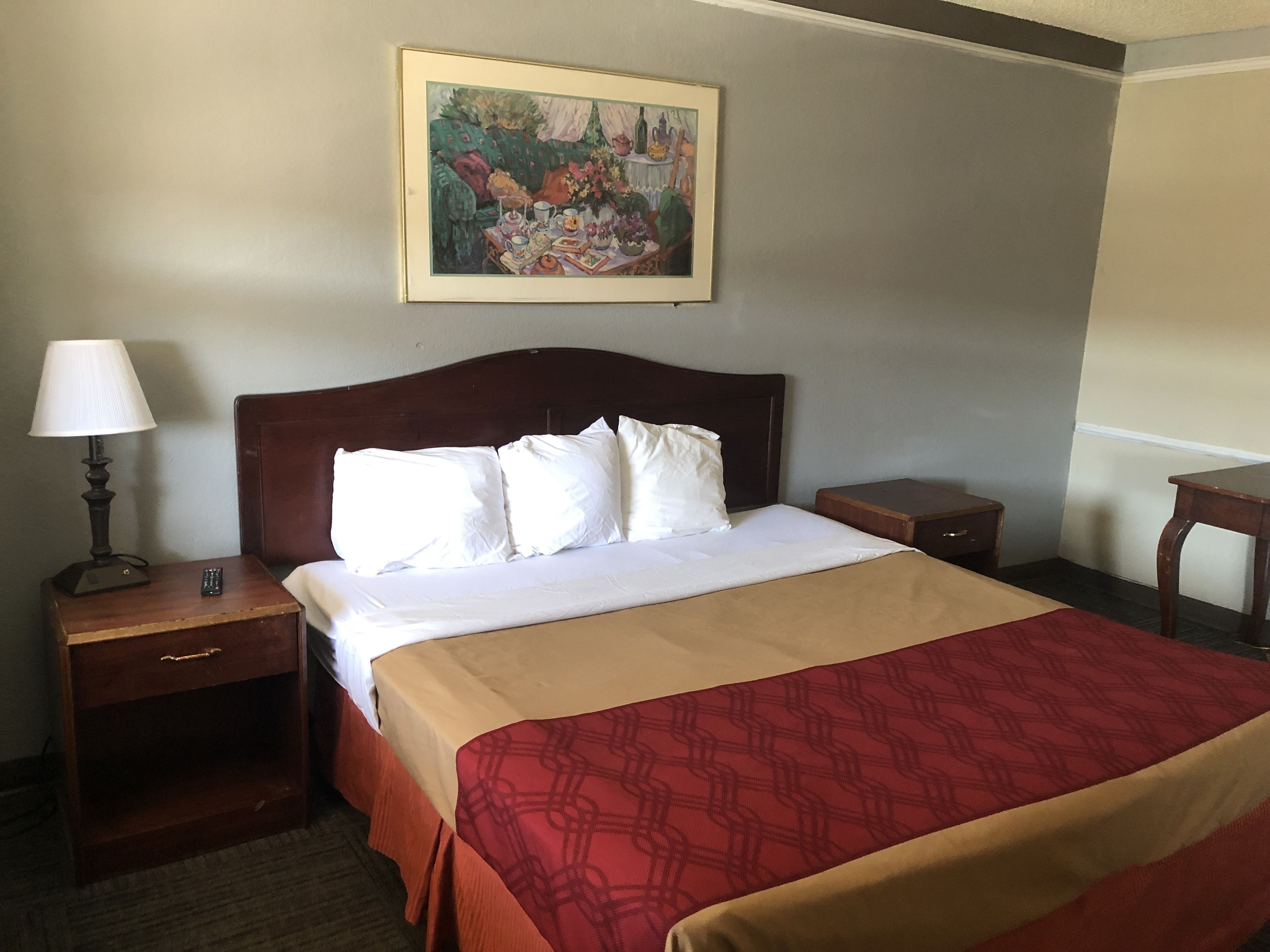 A hotel room with a bed, a nightstand with a lamp, a small table, and a painting on the wall.