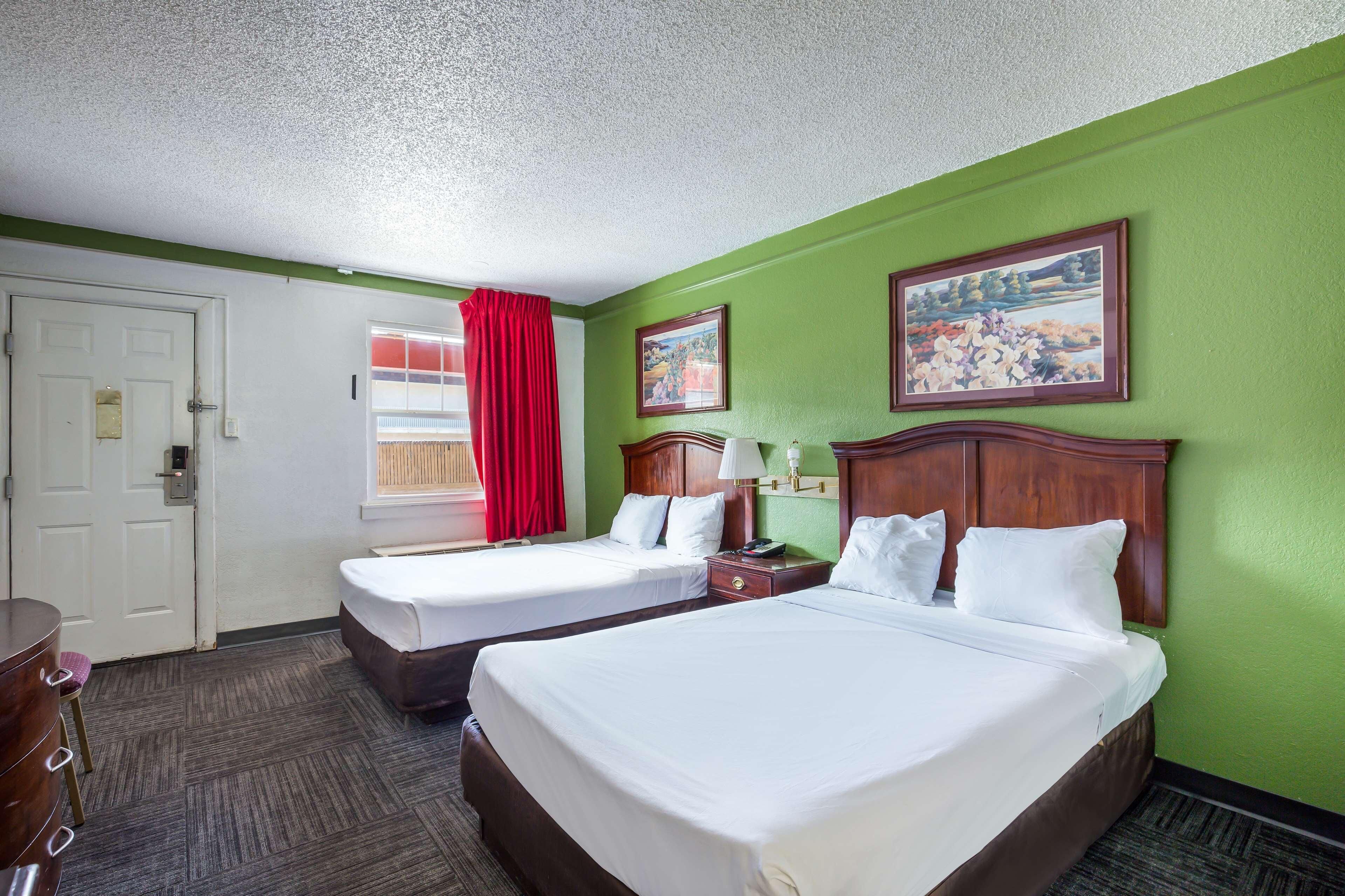 A hotel room with two beds, a desk, a chair, a window with red curtains, and two paintings on the wall.
