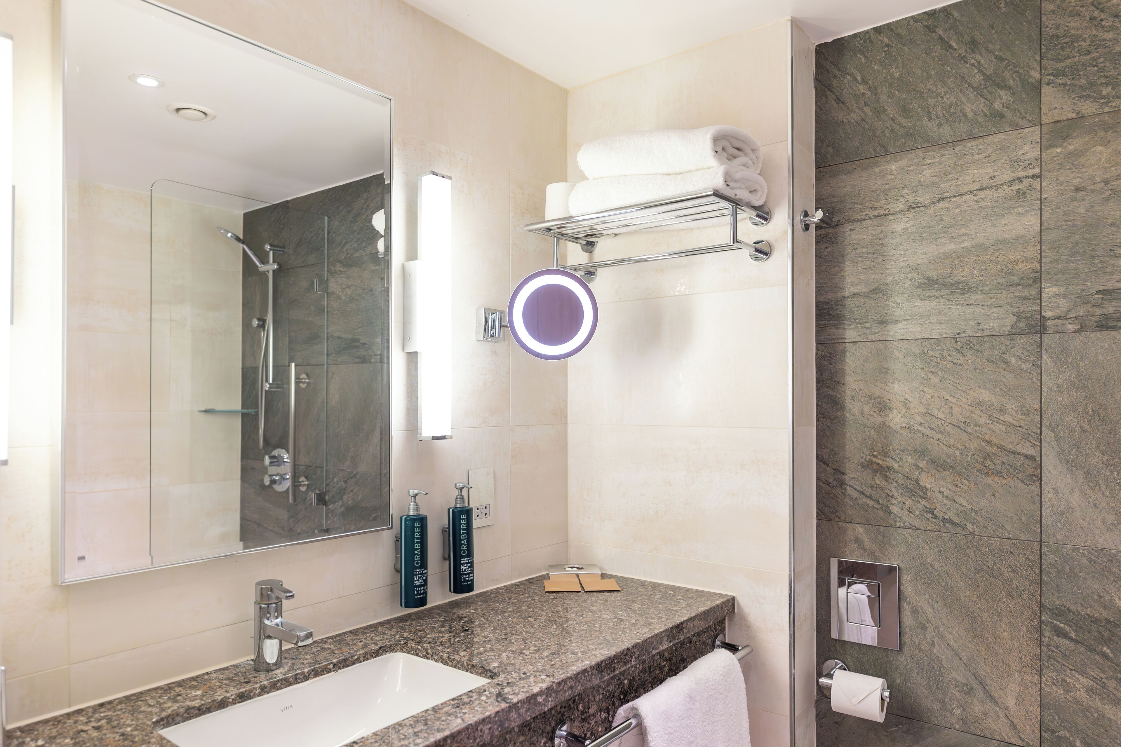 Une salle de bain moderne dotée d’un comptoir en granit, d’un grand miroir, d’une douche à parois de verre et d’un porte-serviettes.