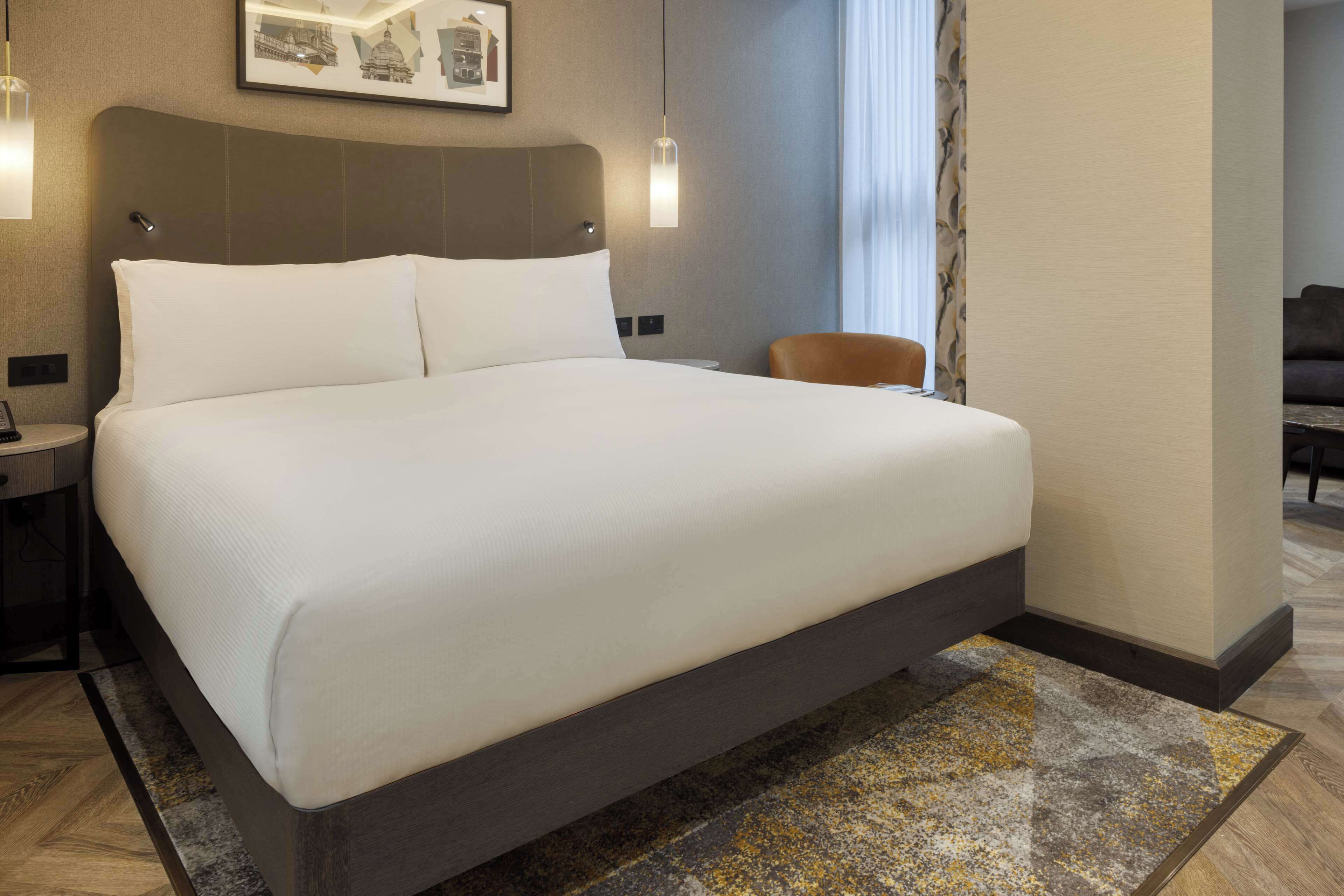 A hotel room with a large bed, a nightstand with a lamp, a chair, and a framed picture on the wall.