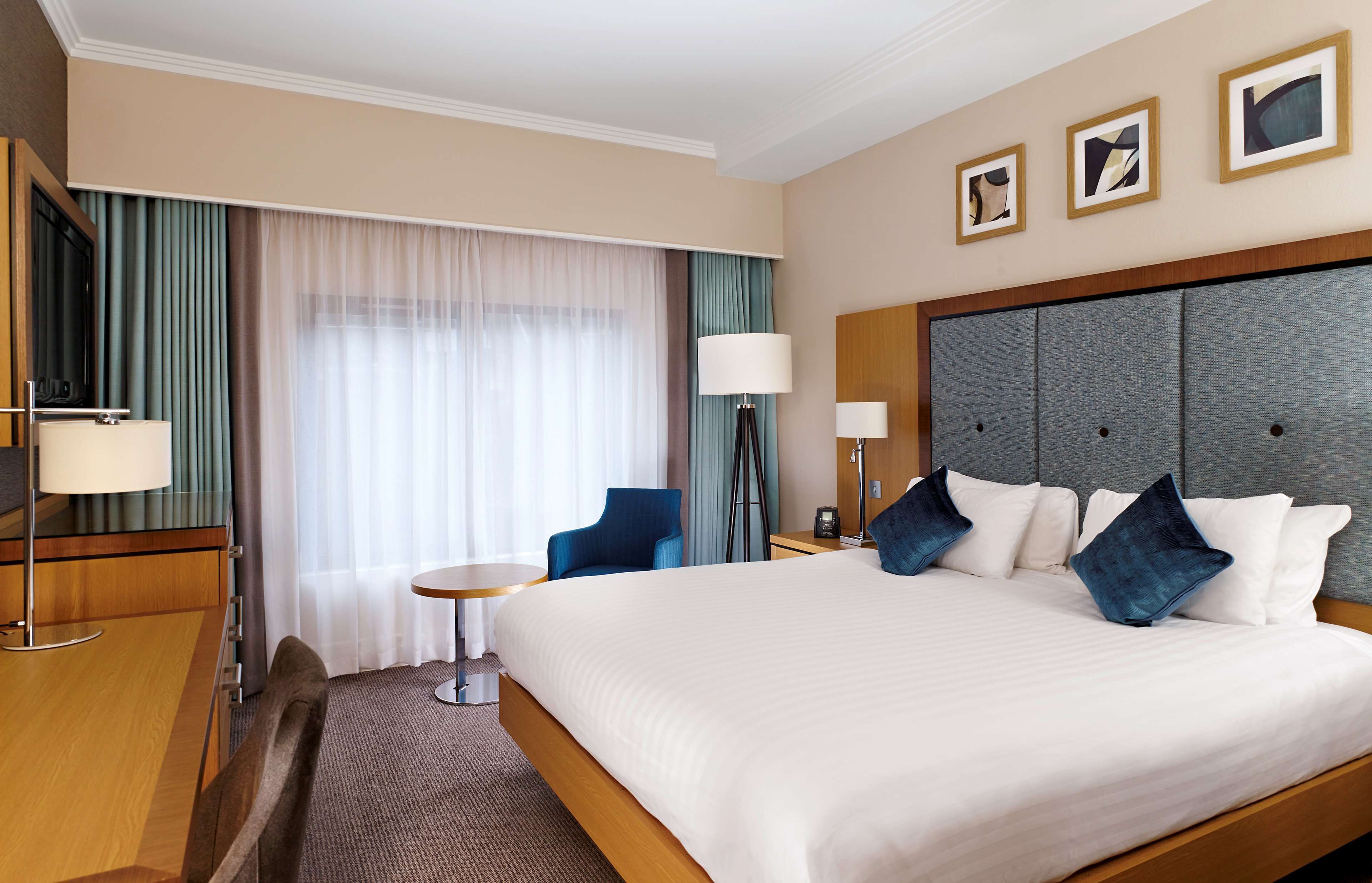 A hotel room with a large bed, a desk with a lamp, a chair, a small table, and a window with curtains.