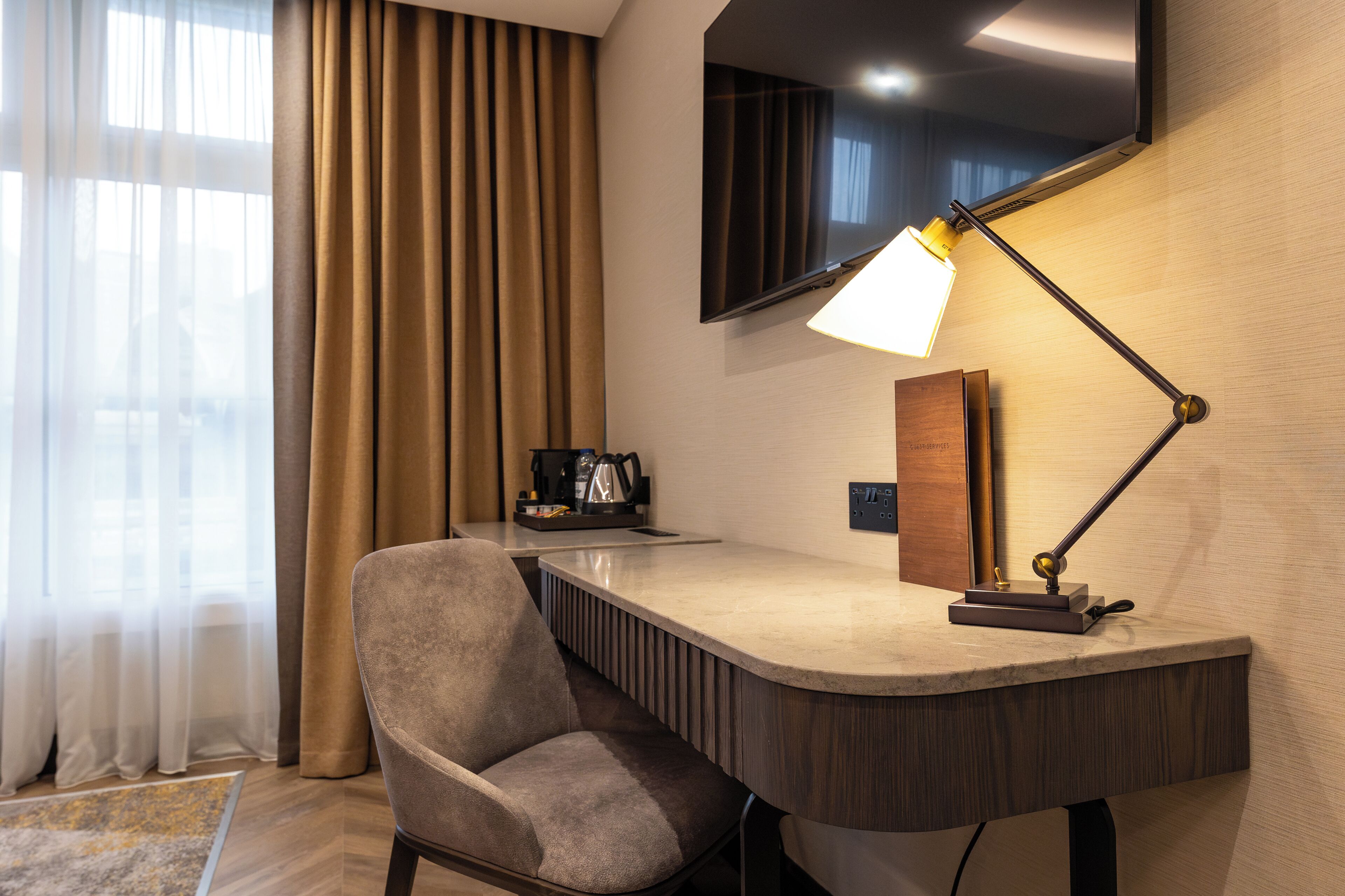 A modern hotel room with a curved desk, a chair, a flat-screen TV mounted on the wall, and a lamp on the desk.