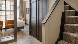 Ein modernes Hotelzimmer mit einem Fischgrätparkett, einem spiegel mit Holzrahmen, einem Bett, einer Sitzecke mit zwei Stühlen und einer Treppe mit strukturiertem Teppich.