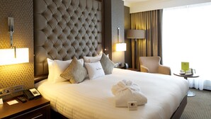 Ein Hotelzimmer mit einem großen Bett, einem gepolsterten Kopfteil, einem Nachttisch mit Lampe, einem Sessel und einem kleinen Tisch mit Tasse und Buch.