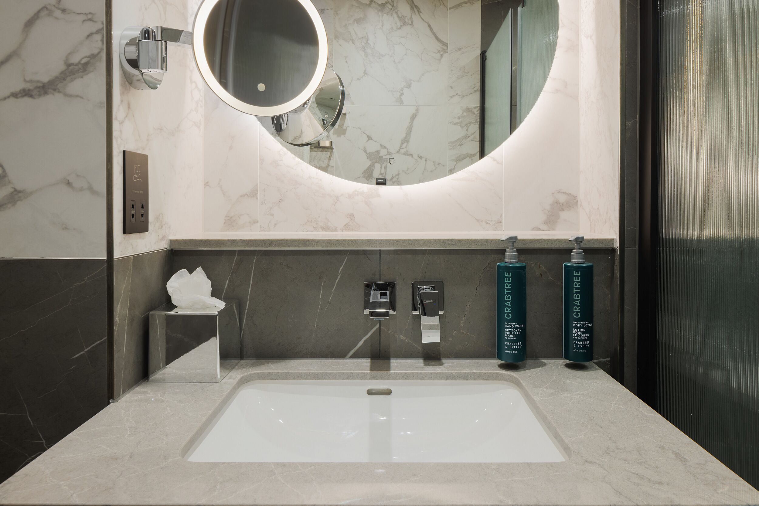 Une salle de bain moderne avec un lavabo en marbre, un miroir rond et deux flacons de lotion pour le corps de la marque Crabtree.