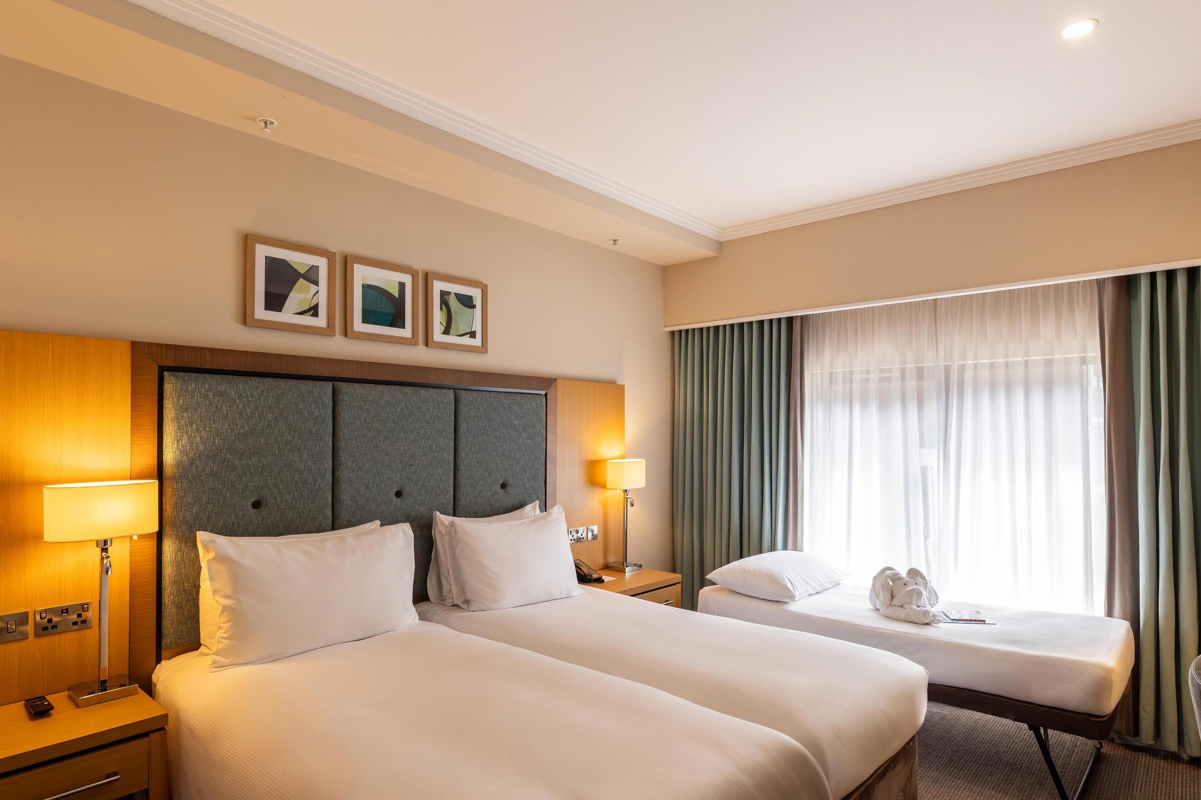 A hotel room with two beds, a headboard, bedside lamps, a nightstand, a window with curtains, and a wall-mounted artwork.