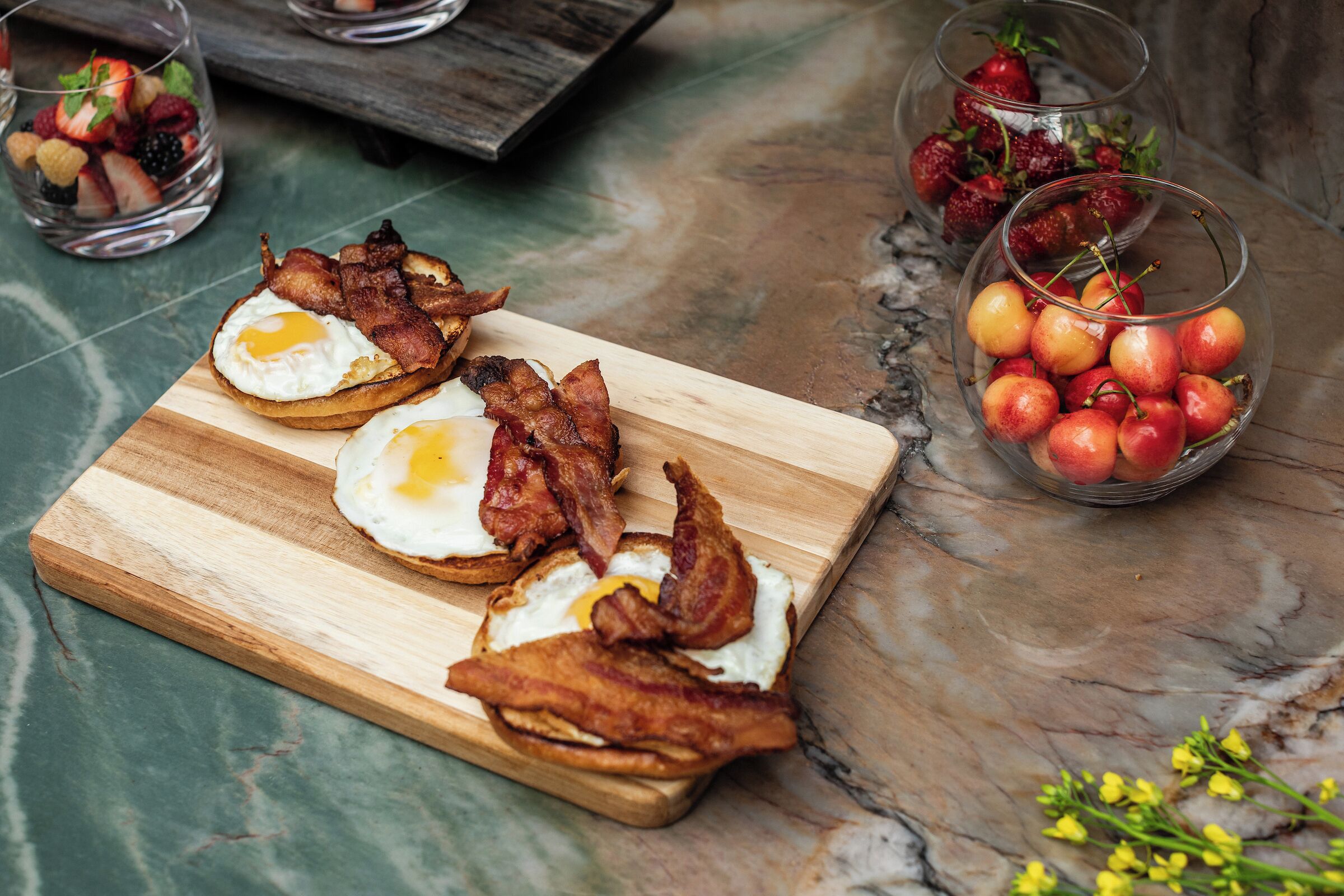 Une planche en bois sur laquelle sont disposés trois morceaux de pain grillé, chacun garni d’un œuf au plat et de lardons, un bol de cerises et un verre de fraises.