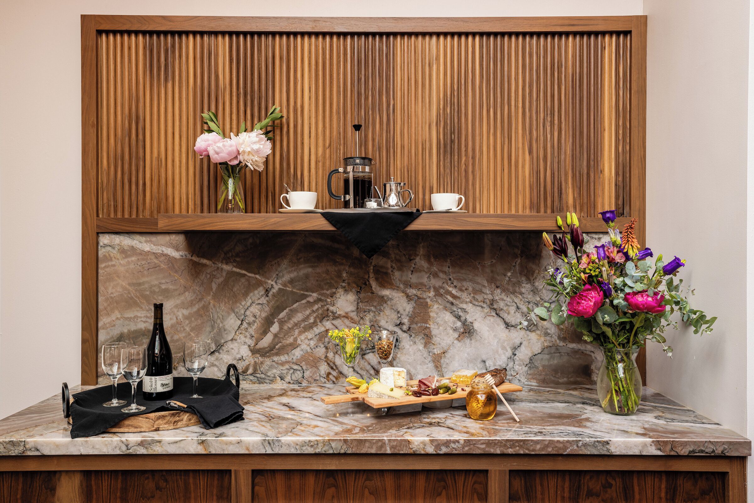 A marble countertop with a coffee maker, two cups, a bottle, a tray with food, and a vase with flowers.