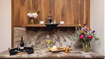 A marble countertop with a coffee maker, two cups, a bottle, a tray with food, and a vase with flowers.