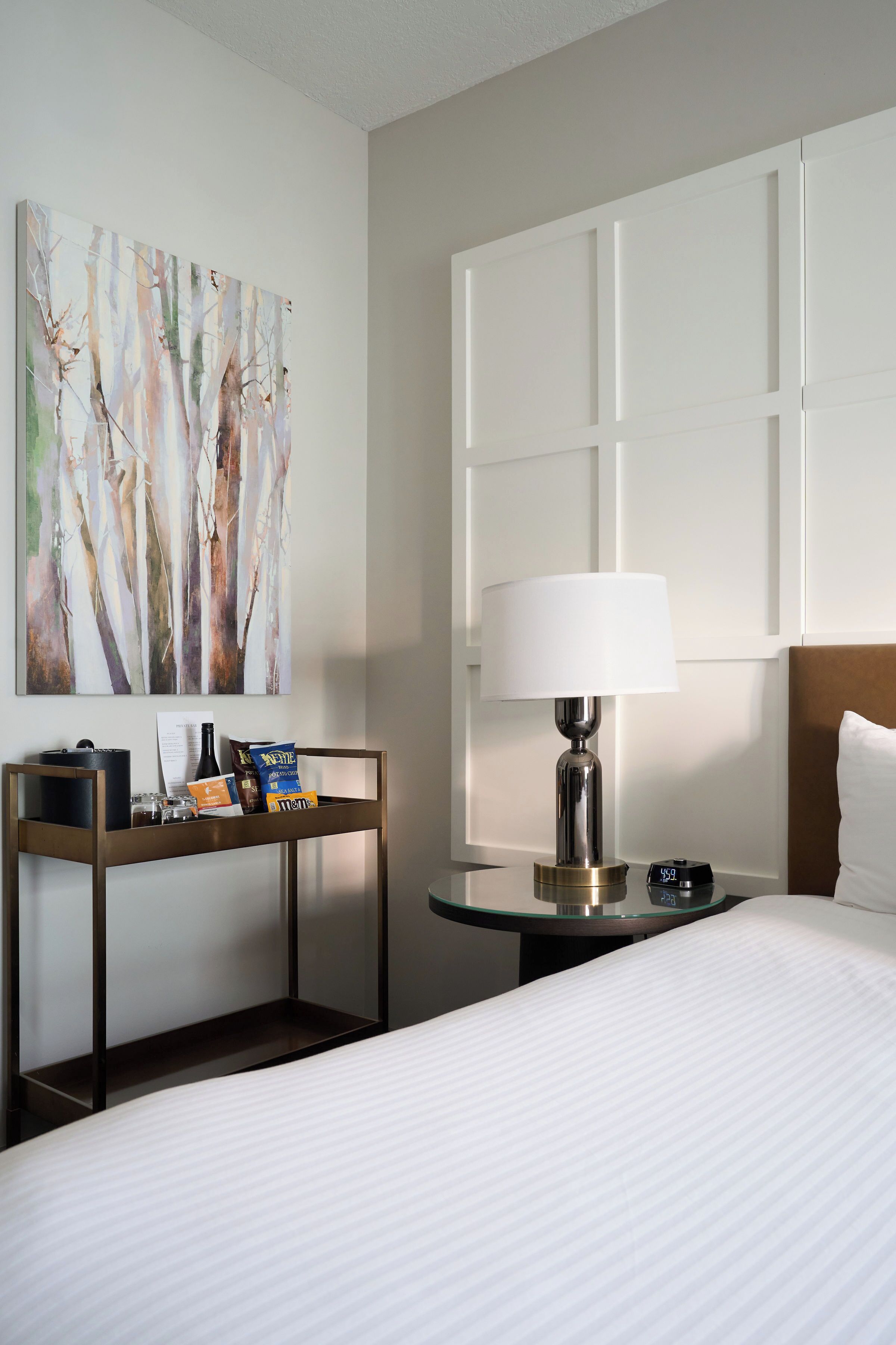 A hotel room with a bed, a bedside table with a lamp, a tray with snacks, and a painting on the wall.