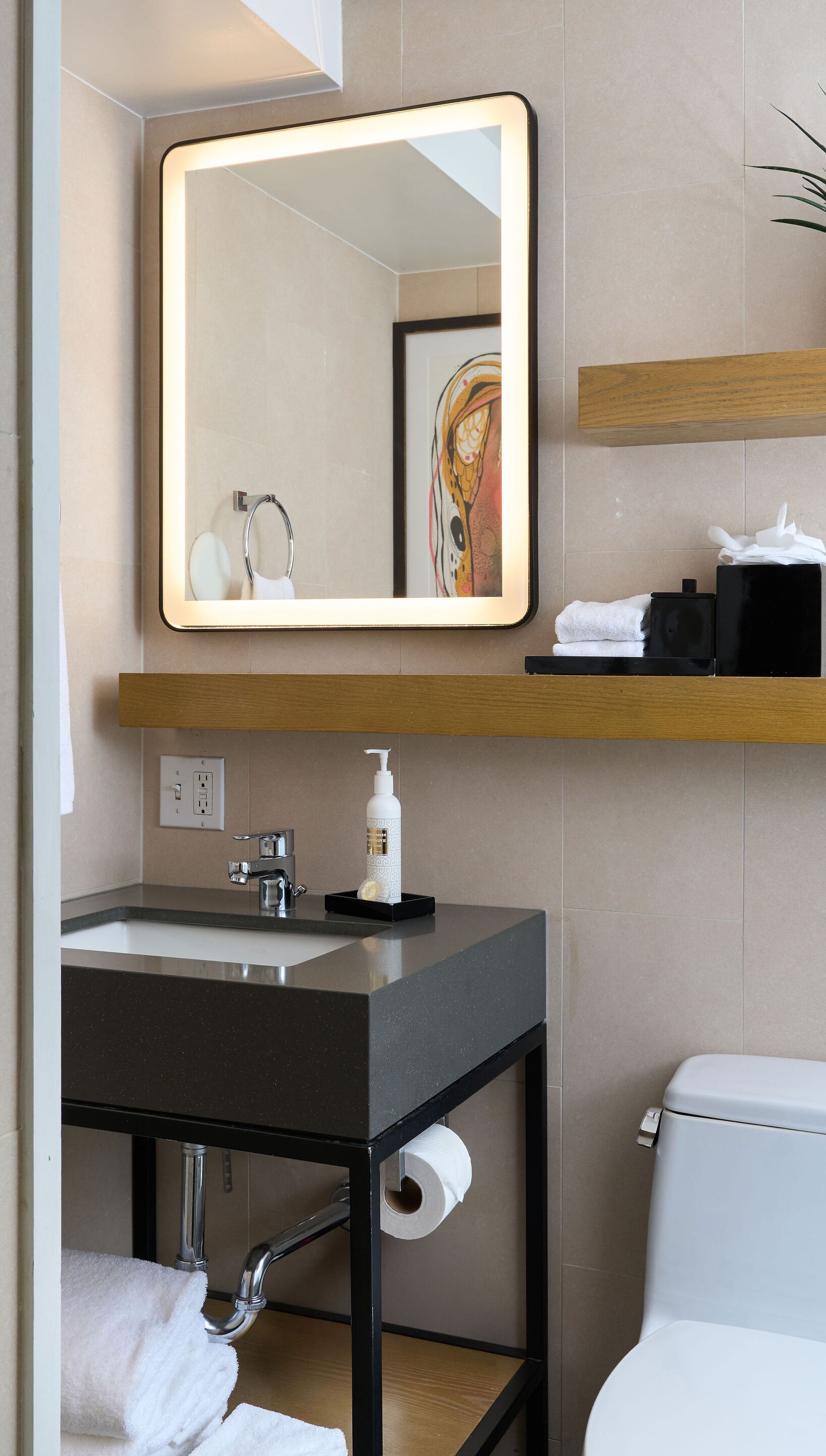 Un baño moderno con un lavabo negro, un espejo con marco iluminado, un inodoro y un perchero para toallas.