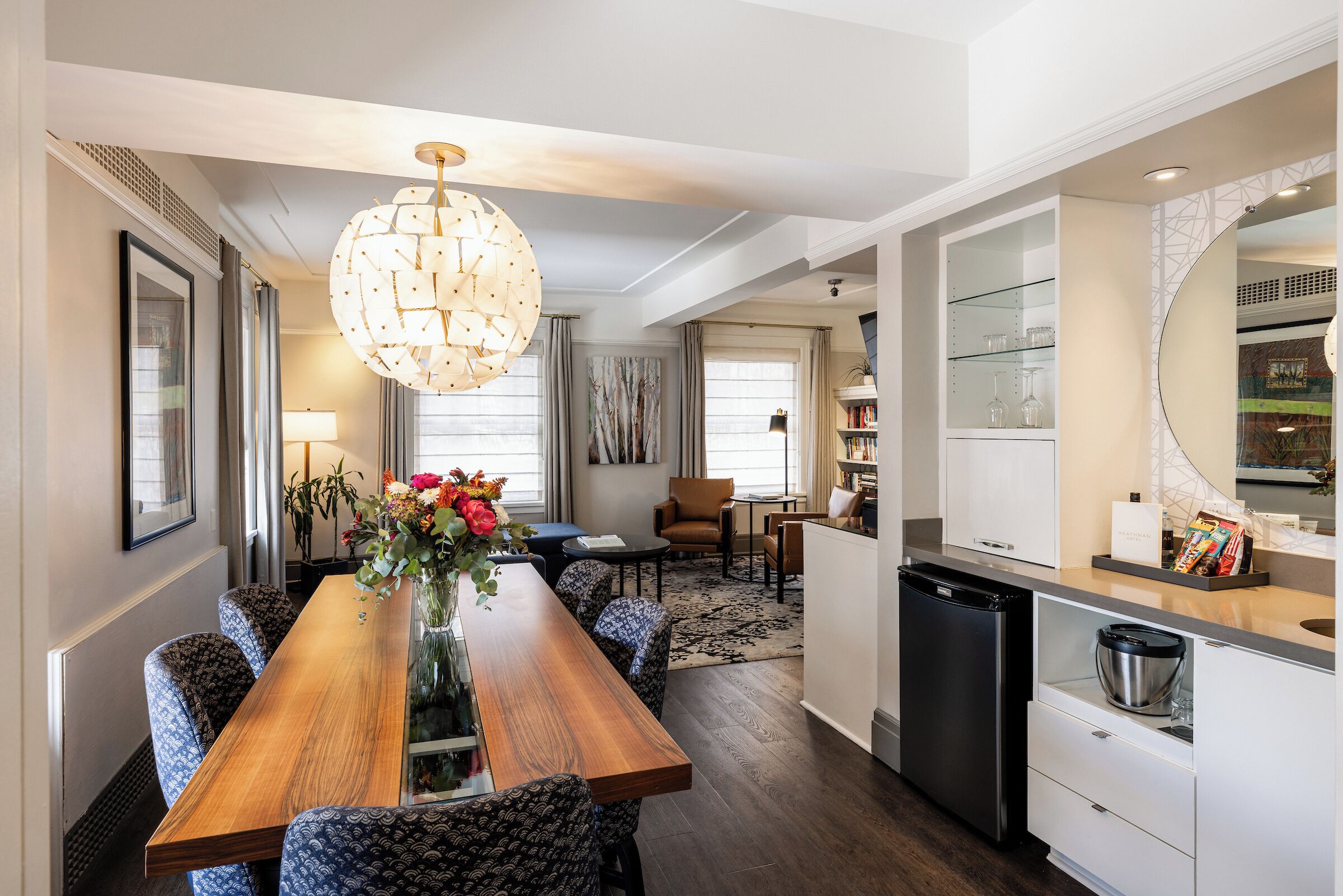 A modern kitchen with a built-in dishwasher, a dining area with a wooden table and patterned chairs, and a large chandelier.