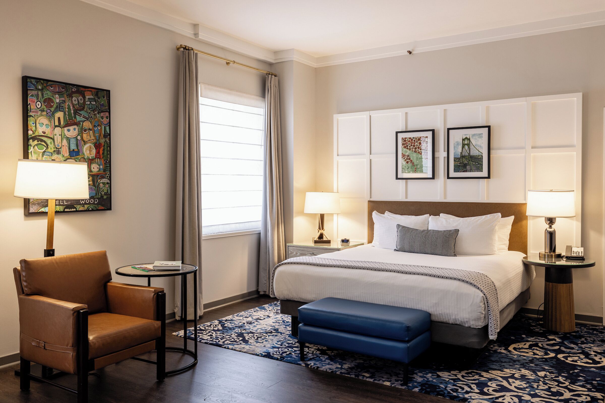 Une chambre d’hôtel avec un lit, deux tables de chevet avec des lampes, une chaise, un pouf bleu, un tapis à motifs et un grand tableau abstrait accroché au mur.