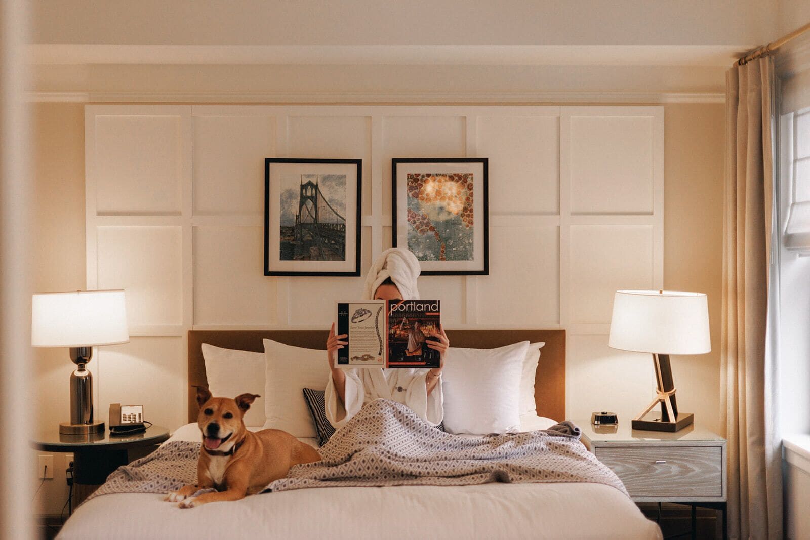 Habitación de hotel con una cama, dos lámparas, un cuadro de un puente y un cuadro de jirafas, además de un perro.