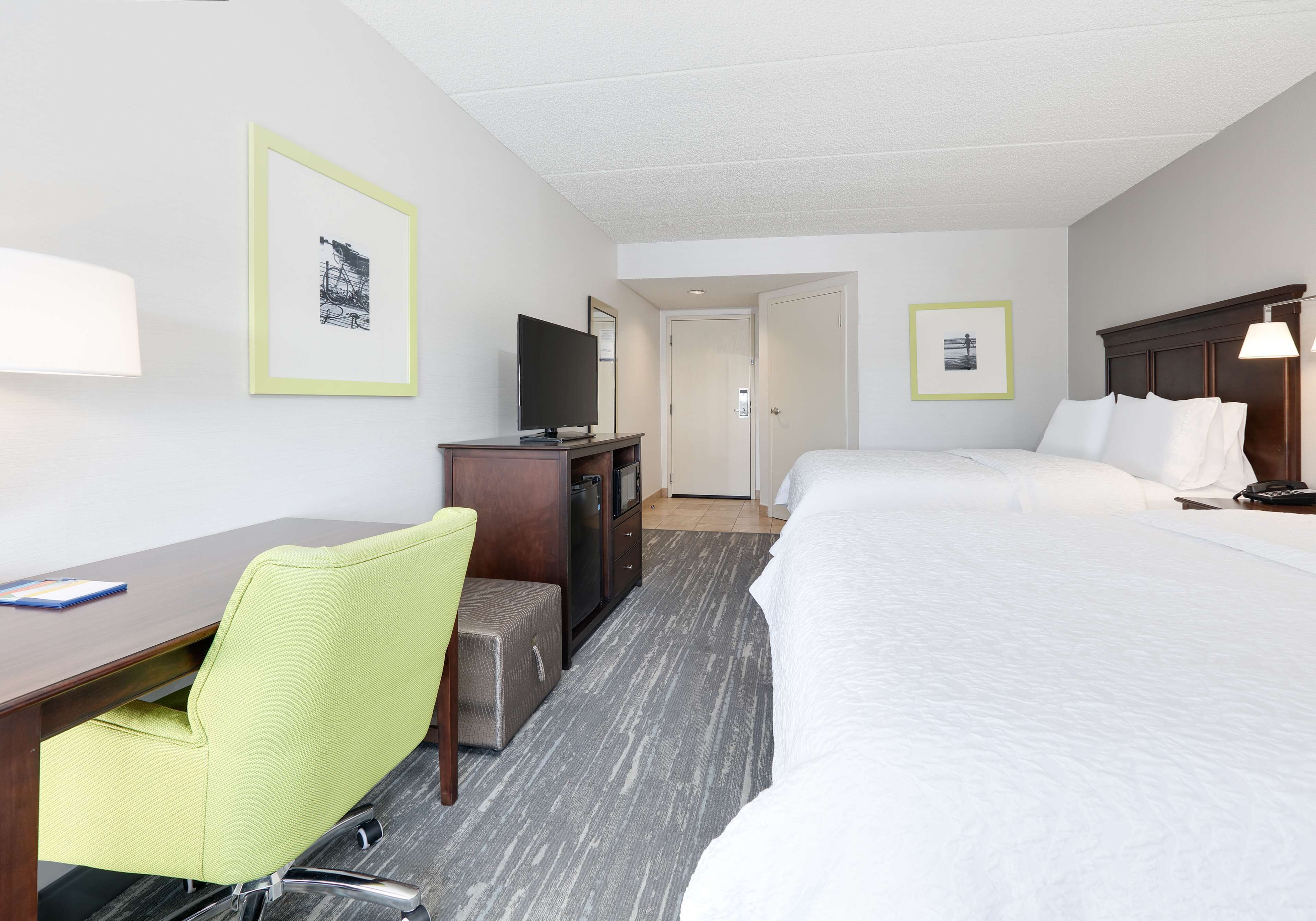 A hotel room with a desk, a green chair, a bed, and a television.