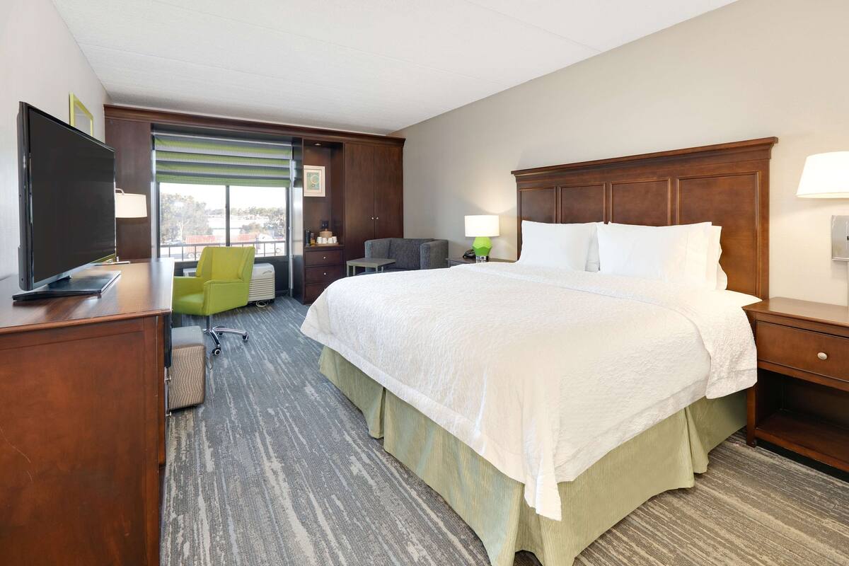 A hotel room with a large bed, a desk with a TV, a chair, and a window with blinds.